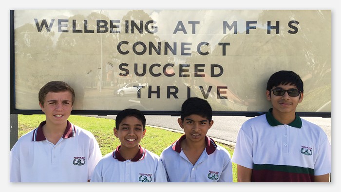 Four high school boys smiling in front of their school sign with the motto Wellbeing at MFHS, Connect, Succeed, Thrive