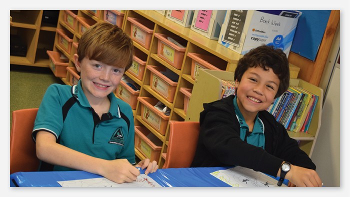 Two Aldavilla public school students sitting at a desk and smiling for the camera.