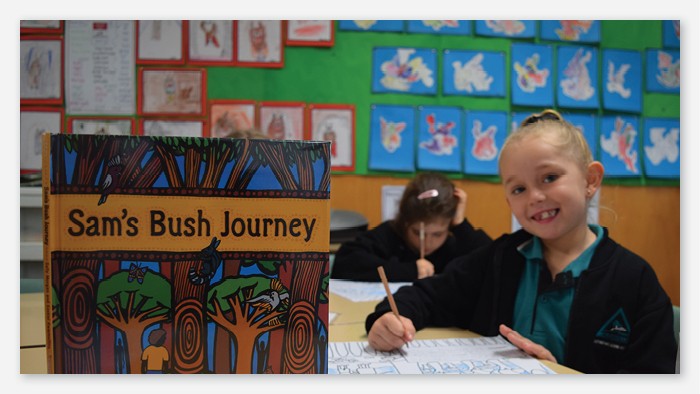 An Aldavilla public school student colouring in with another student in the background. A book titled Sams Bush Journey is in the foreground. The books front cover is an artists painting of a bush scene.