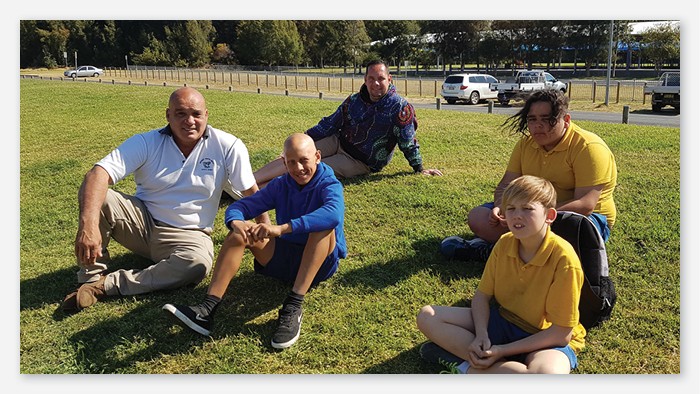 Three Batemans bay public school students and two teachers sitting out in a field watching sport.