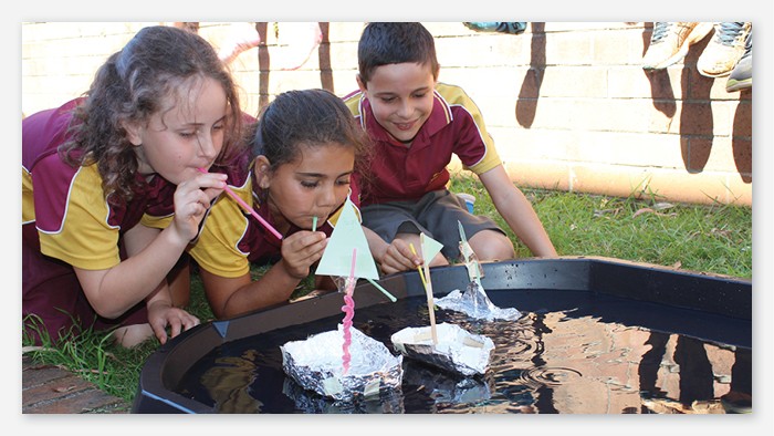 Three Westport public school students blowing into straws to propel tinfoil sailing boats across a small pool.