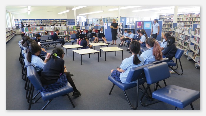 A large group of students sit in a circle of chairs in the school library.