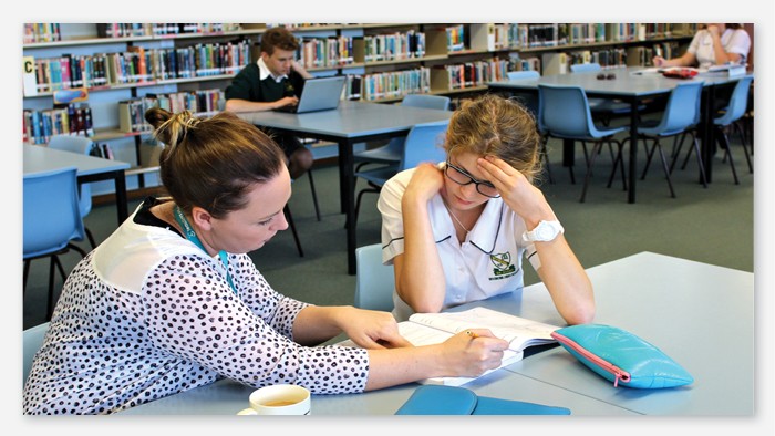 A teacher helps a female student with a writing task in the school library.