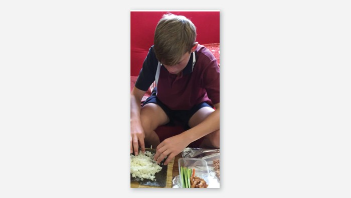 A student sitting down making sushi. He is rolling a sushi roll.