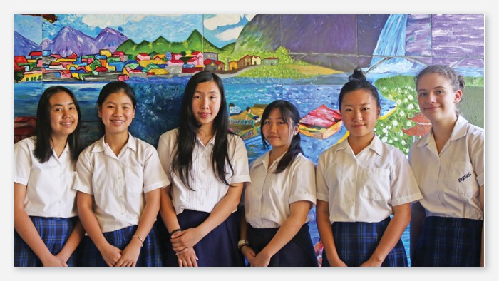 Six students standing in front of a colourful mural depicting a village scene.