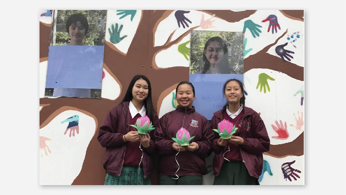 Three students standing in front of a mural holding pink lotus flowers made out of paper.