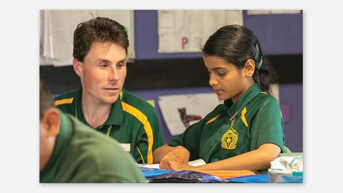 A teacher sits with a young girl as she is doing her school work. They both wear Girraween school colours.