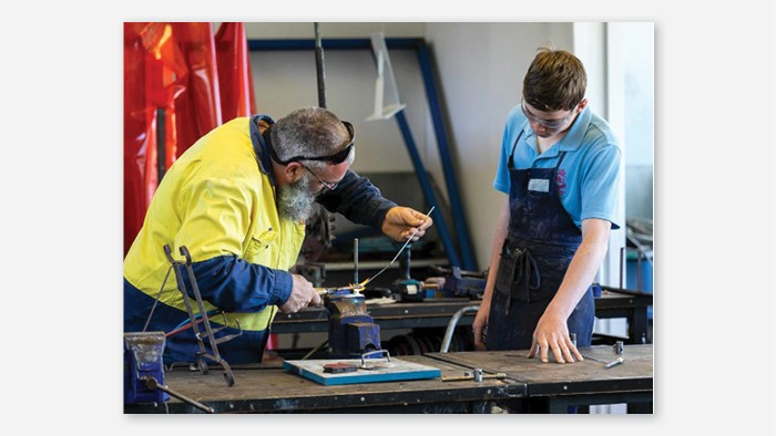 A man and a student in a steel workshop. The man is working on a piece of steel while the student watches intently.