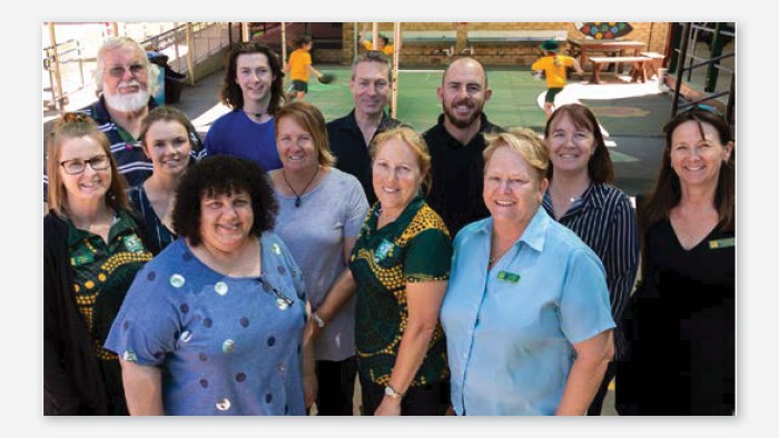 A group of staff members at Wentworth Public School look up and smile at the camera. Some wear shirts in school colours with Indigenous patterns.