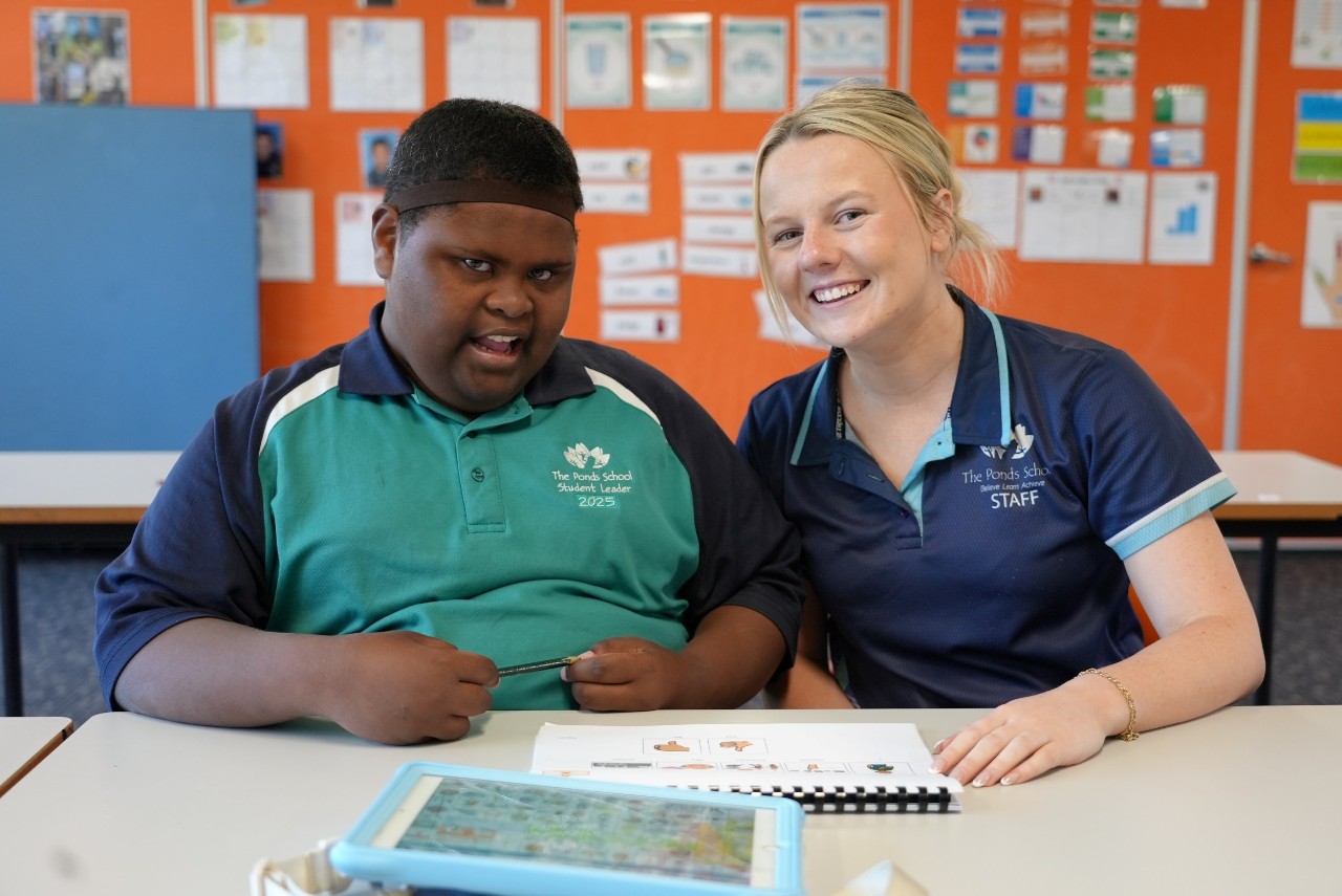 A student and teacher sitting at a desk smiling for the camera