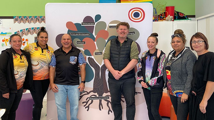 A group of seven smiling people stand in front of a media wall featuring a graphic of a tree in earthy colours. Two raised arms joined at the palms form the trunk of the tree. Black stickers, which feature key themes from the phase 1 report written in white text, are stuck on the leaves of the tree. Four of the people pictured wear polo shirts with brightly coloured Aboriginal artwork.