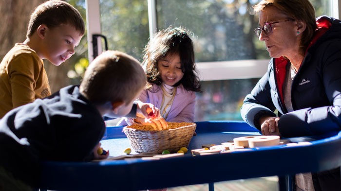 Female educator and 3 young students playing with wooden blocks at an activity table inside an Early Childhood Education centre
