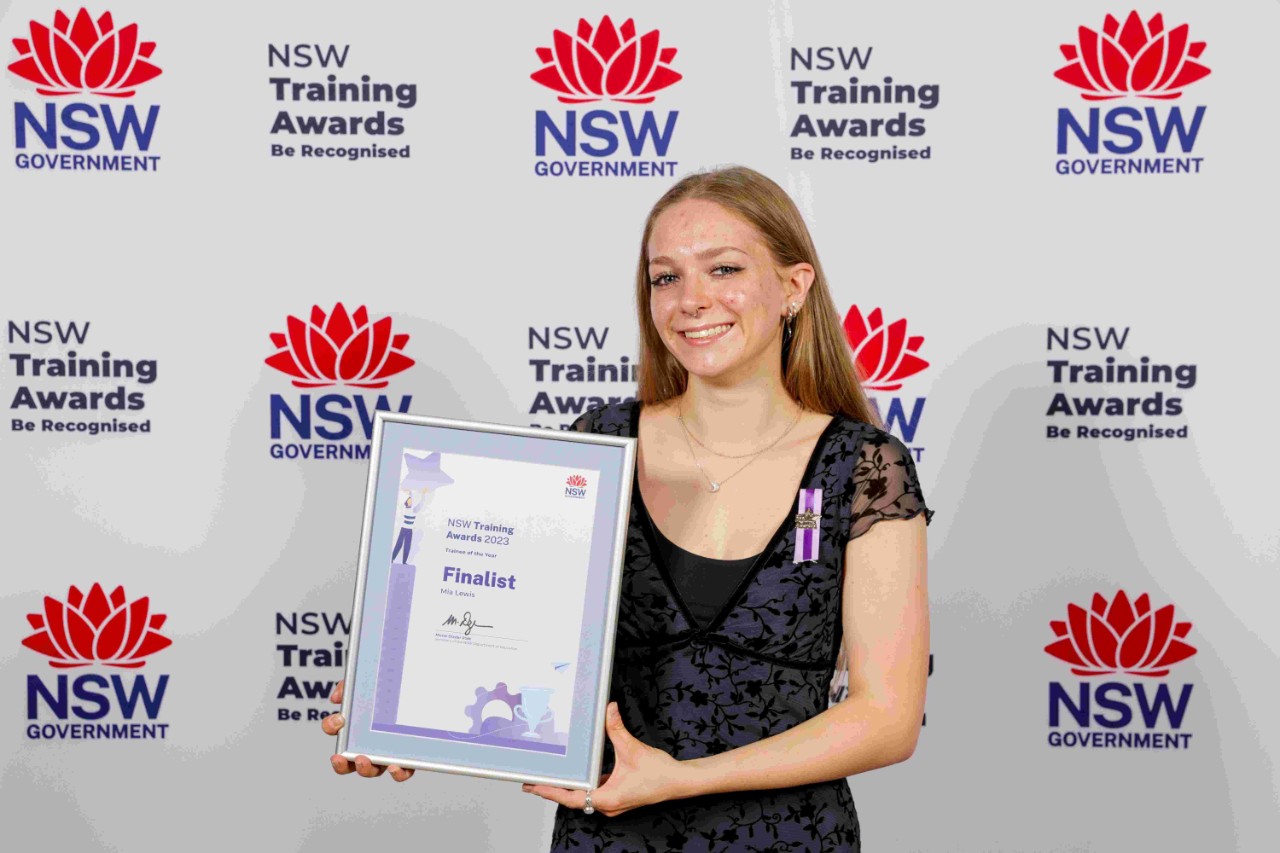 A young woman smiling and holding an award stands in front of a media wall with multiple versions of the red waratah and NSW Government words in blue and the words NSW Training Awards Be Recognised in black.