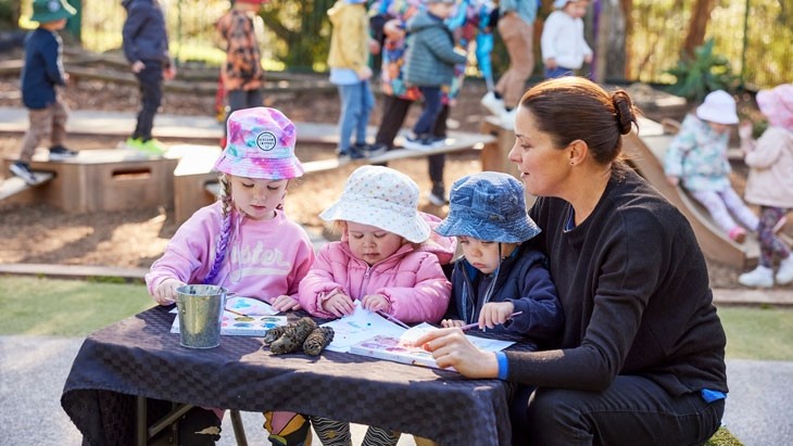 Three young children wearing hats sit next to a female educator at an outdoor table. The children are painting on paper using watercolour paints. Two water colour palettes, a metal paint cup and 3 banksia cones are scattered across the table, which is covered in a black checked tablecloth. In the background, a group of children are playing on outdoor equipment, with an educator in a brightly coloured shirt supervising them.