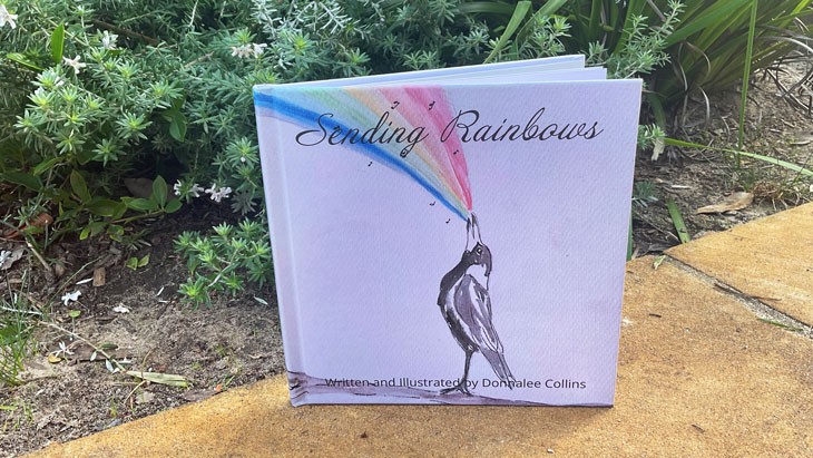 A Sending Rainbows is propped open in an outdoor garden. The cover features watercolour artwork of a magpie standing on the ground with its beak raised to the sky with a rainbow flowing out. The Written and illustrated by Donnalee Collins runs along the bottom of the cover.