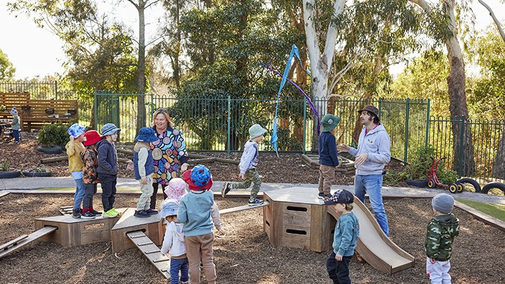 A group of young children play on wooden play equipment outside, with different platforms and balance beams. Two educators are interacting with the children as they play.