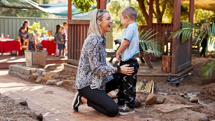 A woman with shoulder length blonde hair, who wears a snakeskin-print blouse and black trousers, crouches down in front of her son. He is wearing a light blue t-shirt and long dark pants. They are both smiling. The mother and son are standing outside in front of a timber gazebo. An educator and group of children are visible in the background.