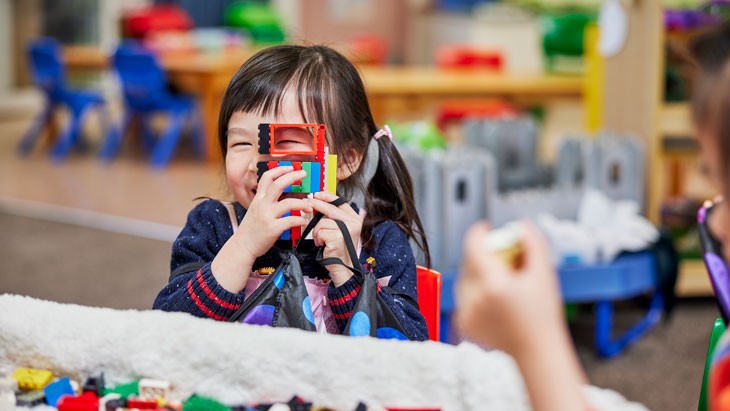 A young girl with dark brown hair in pigtails holds a small structure made from colourful building bricks in front of her face, she is peering through one of the gaps and smiling. Brightly coloured furniture and equipment is visible behind her.