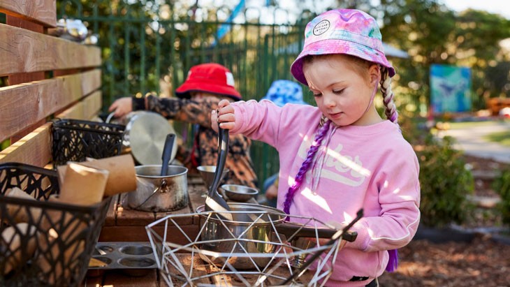 A child plays in an outdoor kitchen holding a black plastic spoon which she is using to stir items in a metal saucepan