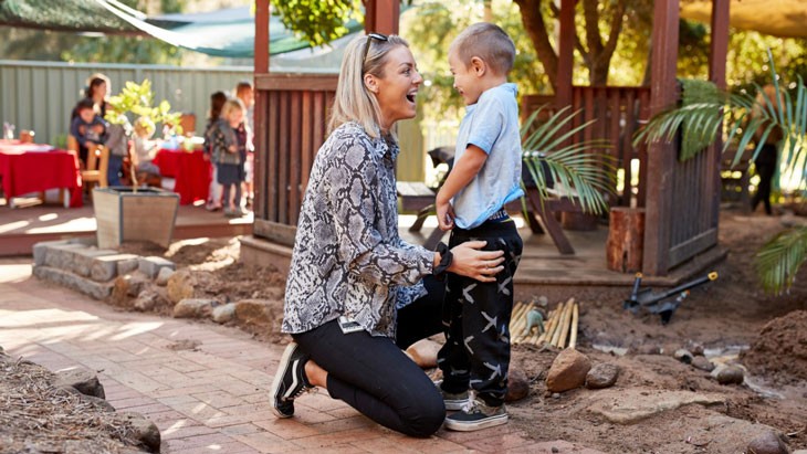 A smiling woman kneels on the ground in front of a young child. They are standing in an outdoor area. An educator with a group of children is visible in the background.