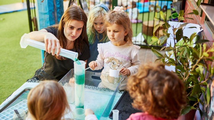 A group of children and a female educator sit around a table. They are engaging in a science experiment, with the female educator pouring green liquid into a beaker.