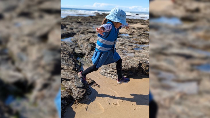 A young child steps off a rock onto sand at the beach. Waves are crashing in the background.