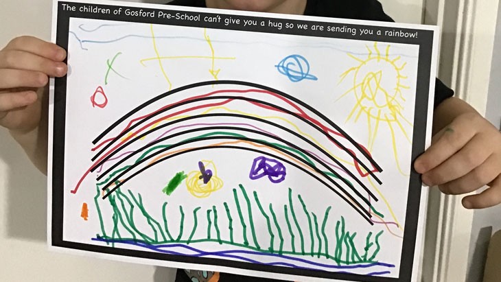 A young child holds a white piece of paper. An outline of a rainbow is in the middle of the page, which has been coloured in with marker pens.