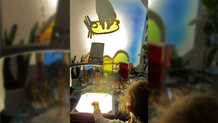 A child positions a cut-out drawing on the glass plate of an overhead projector. An image of a bright yellow creature outlined in black is projected on the wall in front of the child. Colourful props are scattered across the table in front of them.