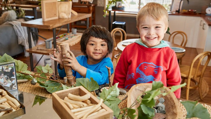 Two happy young children stand together in front of a table covered in a natural fibre mat, with a mirrored panel placed in the centre. Wooden trays filled with loose parts, including twigs and stones, are scattered across the surface. The child on the left is using both hands to stack small offcuts of tree branches on top of each other.