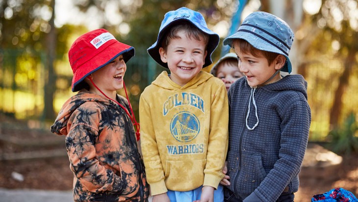 Three smiling young male children wearing bucket hats and hooded jumpers stand together in a leafy outdoor play area. The 2 children on either end face the child standing between them. A shorter child stands behind the children and looks at the camera over their shoulders.