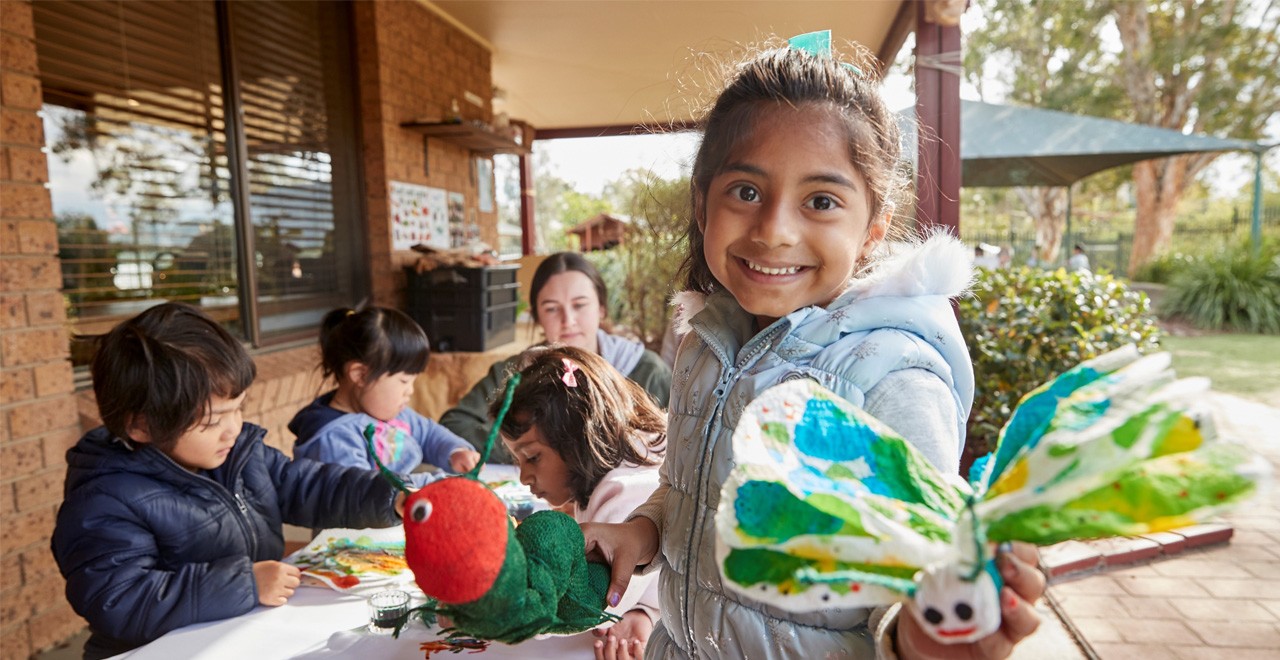 A girl is smiling and holding a paper butterfly outdoors. Behind her, children and an educator are sitting at a crafts table.