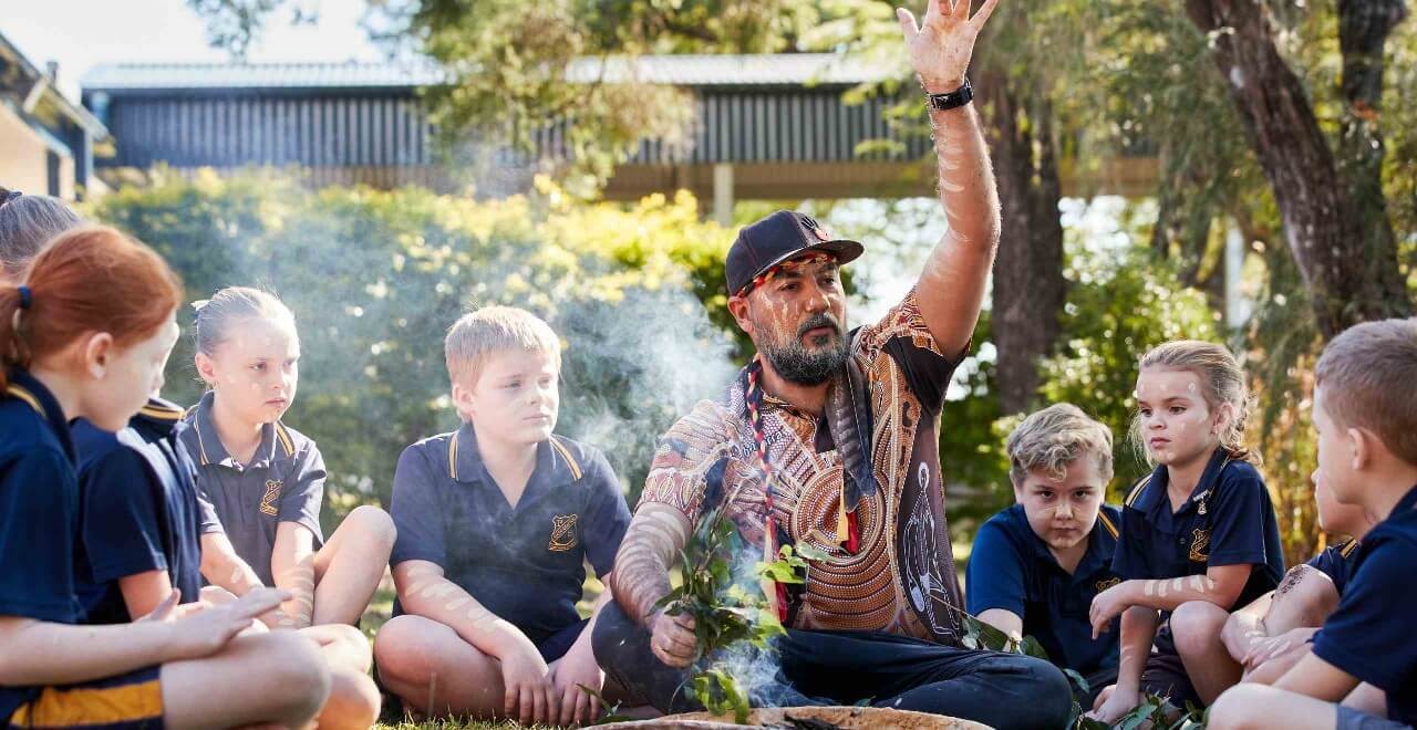 Primary students sitting in a circle with an Aboriginal elder demonstrating a smoking ceremony.