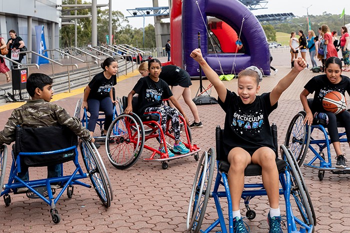 A group of students roll around in wheelchairs designed for basketball on the Qudos Bank Arena forecourt. One student reaches both hands for the sky in a gesture or triumphal exuberance