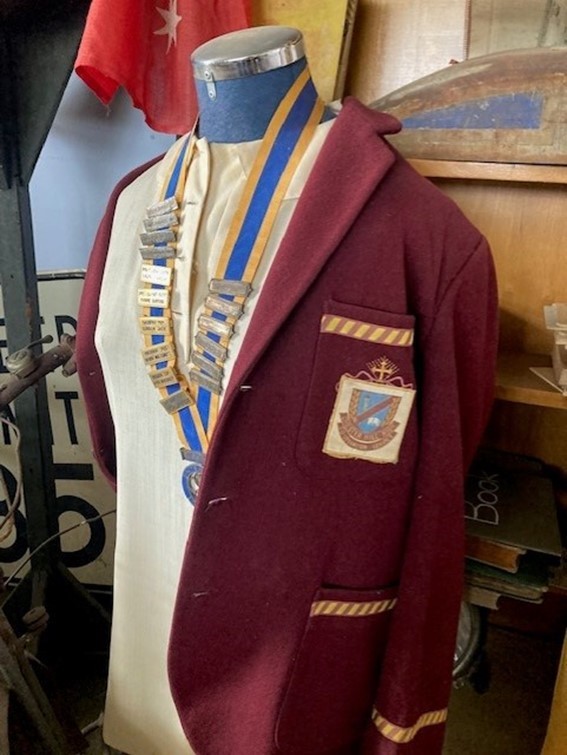 A mannequin wears a faded and dusty old school uniform, along with a medal covered in rectangular pins. These pins are illegible but they show 16 years worth of SRC badges. The uniform consists of a maroon blazer and yellow striped trims at the breast pocket and wrist. The breast pocket shows a religious style logo that has been covered with a sewed on logo from Chester Hill High School.