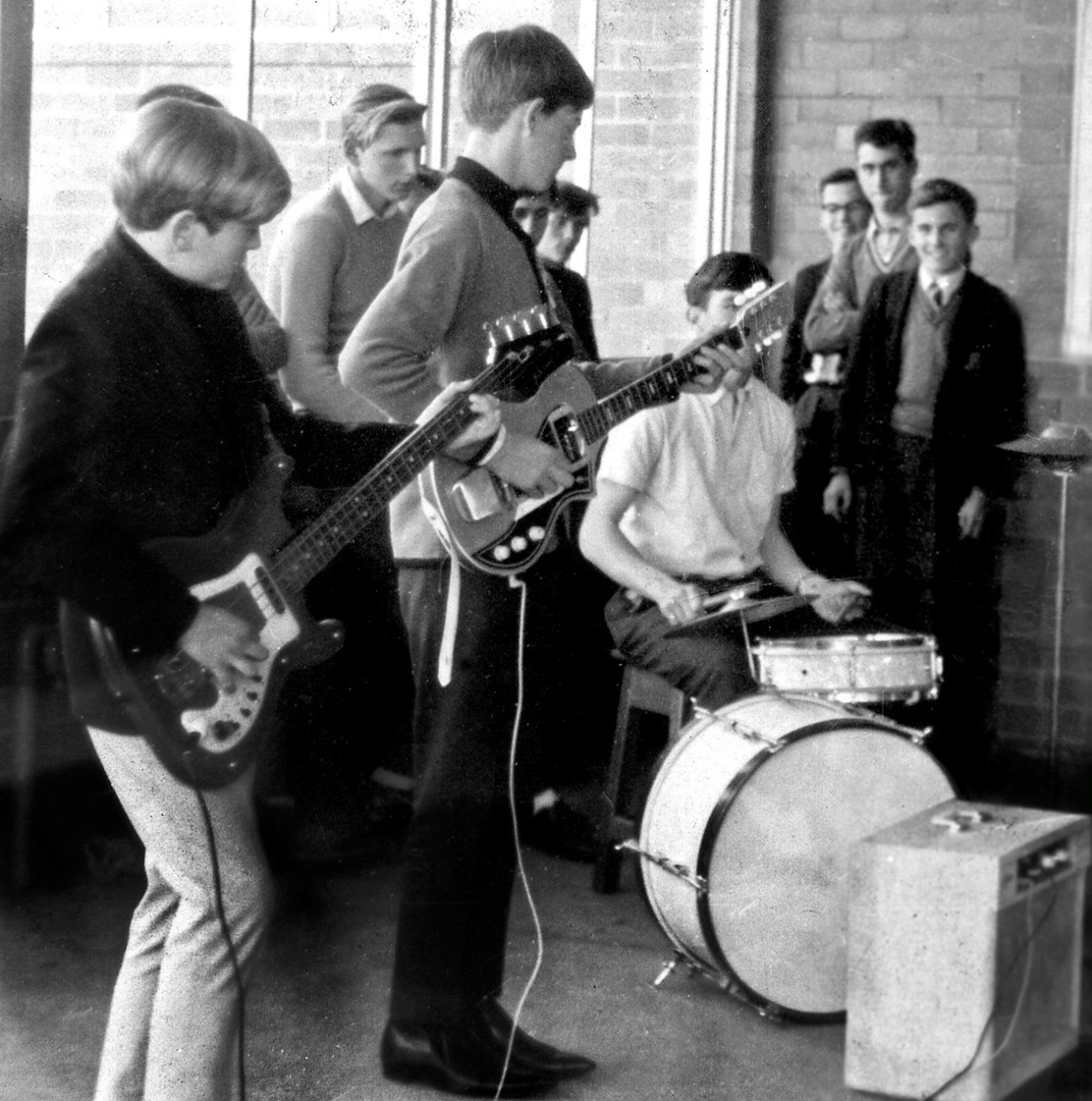 A black and white photograph of the CHHS band playing a small concert at school in 1964. Male bassist and male guitarist are in the foreground with drummer in the middle ground. 8 male students in school uniform are in the background.