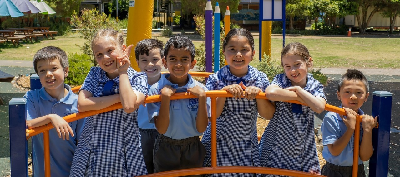 Seven school children smile on a piece of orange play equipment on a sunny day.