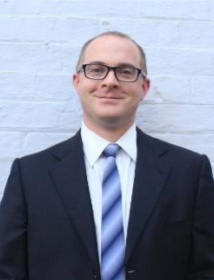 A photograph of Dr Ben Jensen from the torso up. He wears a suit, blue striped tie and glasses and stands in front of a painted brick wall.