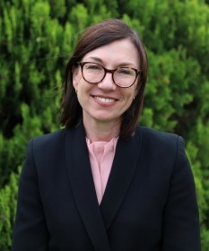 A photo of Mary Doolan, wearing a black jacket, pink shirt and glasses. She is smiling at the camera.