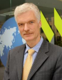 Professor Andreas Schleicher stands in front of a glass wall with an image of the world. He is wearing a suit and tie.