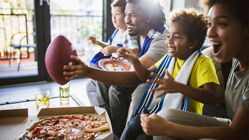 Family sitting on couch, holding football with pizza box in front of them