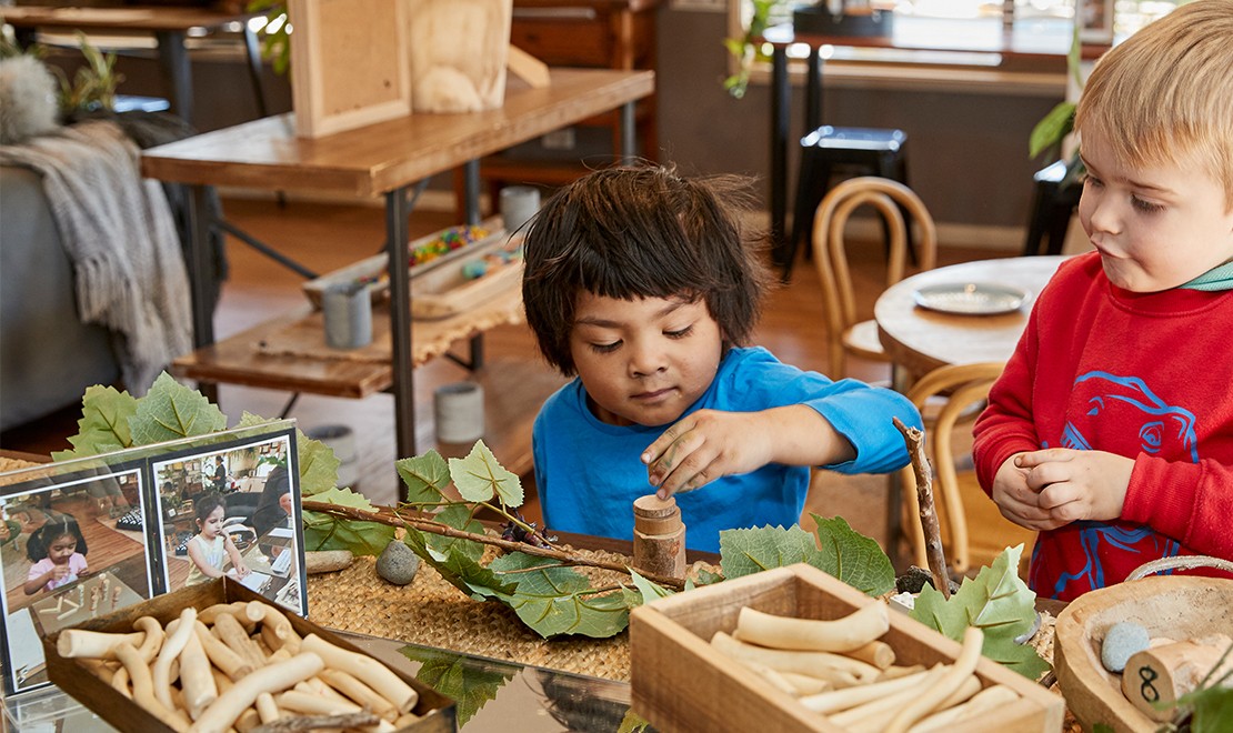 Two young children playing with wood, sticks and leaves at a desk.