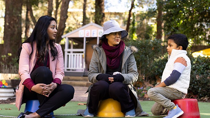 Two female educators and a young male student sitting and talking in outdoor play area of an early childhood education centre.