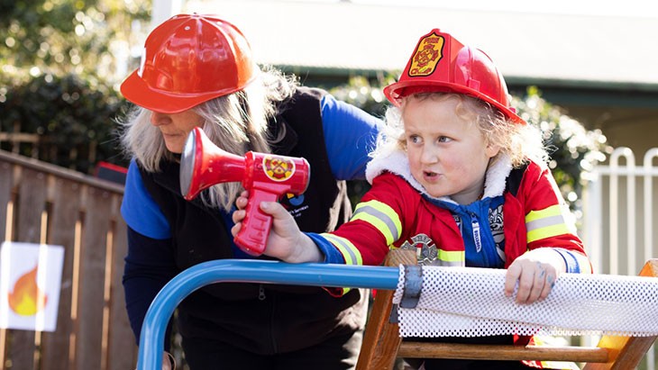 A young female student wearing a firefighter costume stands on outdoor climbing equipment, holding a toy megaphone in her right hand. An educator wearing a red hard hat stands behind her. In the background an illustration of a flame is affixed to a wooden fence.