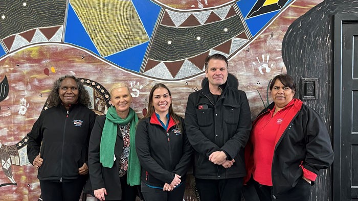 Five Aboriginal Families as Teachers providers and Department of Education staff stand in front of a mural of Aboriginal art.