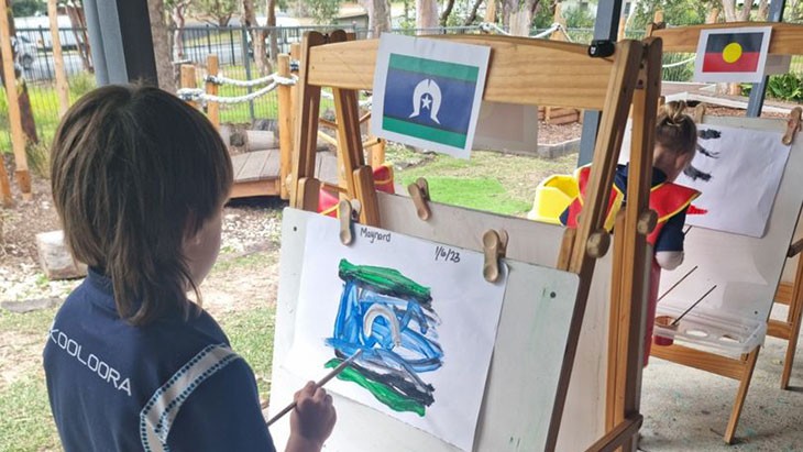A child with brown hair wearing dark blue polo shirt stands at a wooden easel outside painting the Torres Strait Islander flag. A picture of the flag is attached to the top of the easel. Another child wearing a dark blue shirt and red apron painting the Aboriginal flag is pictured in the background.