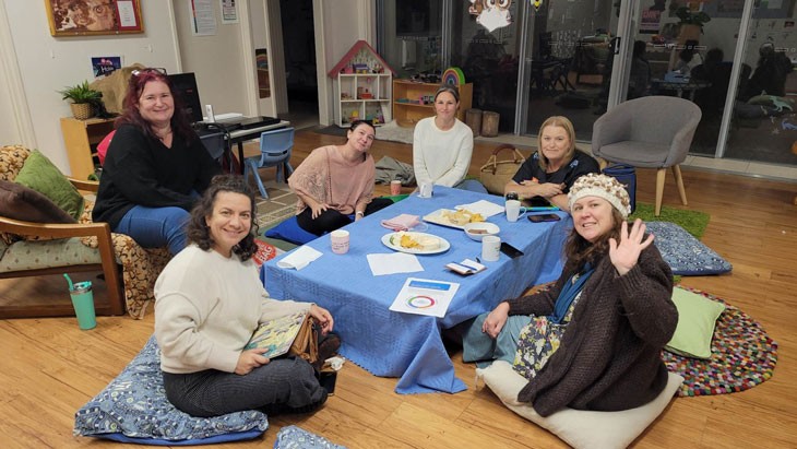 6 women sit on the floor around a low table with food and printed pieces of paper on the table.