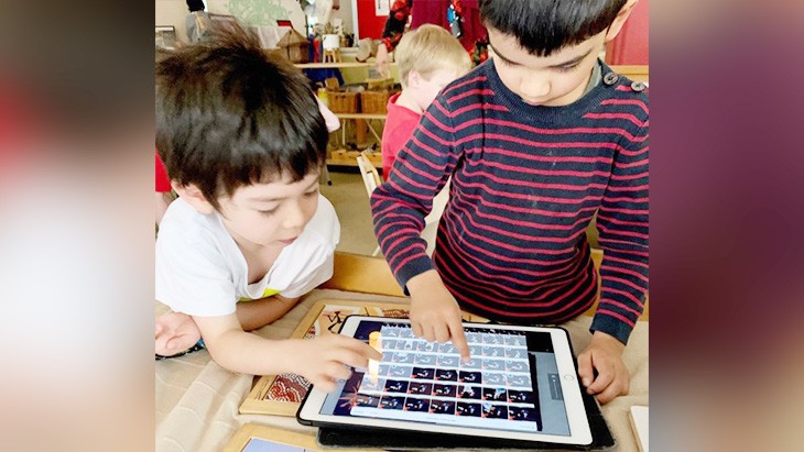 2 children with short dark brown hair, one wearing a white t-shirt and the other wearing a red and black striped sweater, gather around a table using a stop motion app on an iPad.