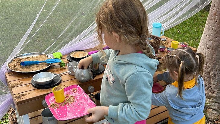 Two young girls seen from the back playing with toy cooking utensils at a table.