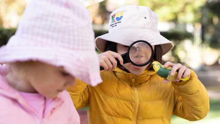 Young female student with another female student in an outdoor area of an early childhood education centre looking through a magnifying glass at a toy budgerigar bird
