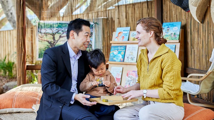 A male guardian in corporate attire sits with a female educator, who wears a yellow sweater, in an outdoor reading area. Both adults are laughing. A young child sits on the mans lap and holds a toy tool. A bookshelf filled with colourful childrens books, floor cushions and a colourful tree artwork and visible in the background.